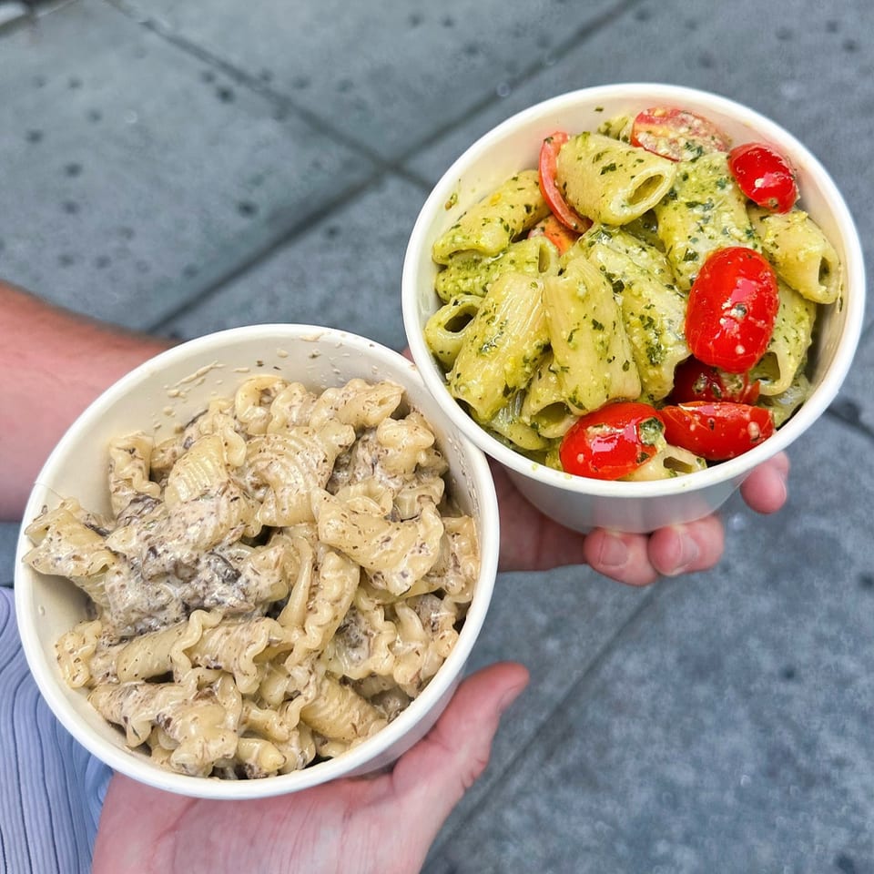 Two hands hold two bowls of Pasta Lab NYC pasta, one pesto with tomatoes, one creamy.