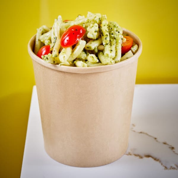 Pesto pasta with cherry tomatoes in a brown paper cup, on a white marble surface.