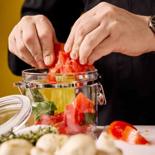 Hands adding diced tomatoes to a glass jar with olive oil and basil for an Italian dish.