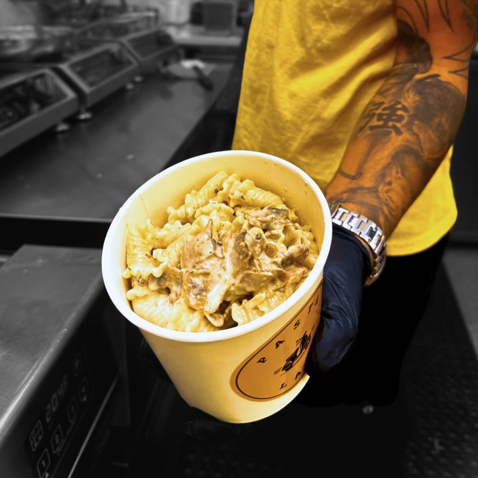 A person in a yellow shirt and black glove holds a cup of creamy Pasta Lab NYC pasta.